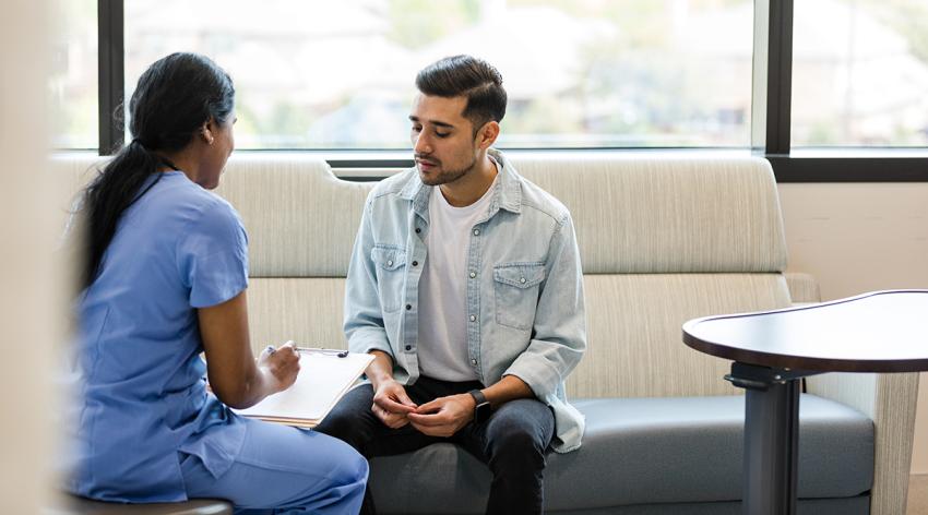 The young adult male sits on the hospital room couch to talk with the hospital insurance specialist to update medical information.