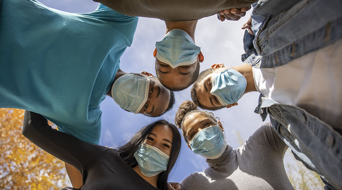 A diverse group of people wearing masks stand in a circle looking down at the camera