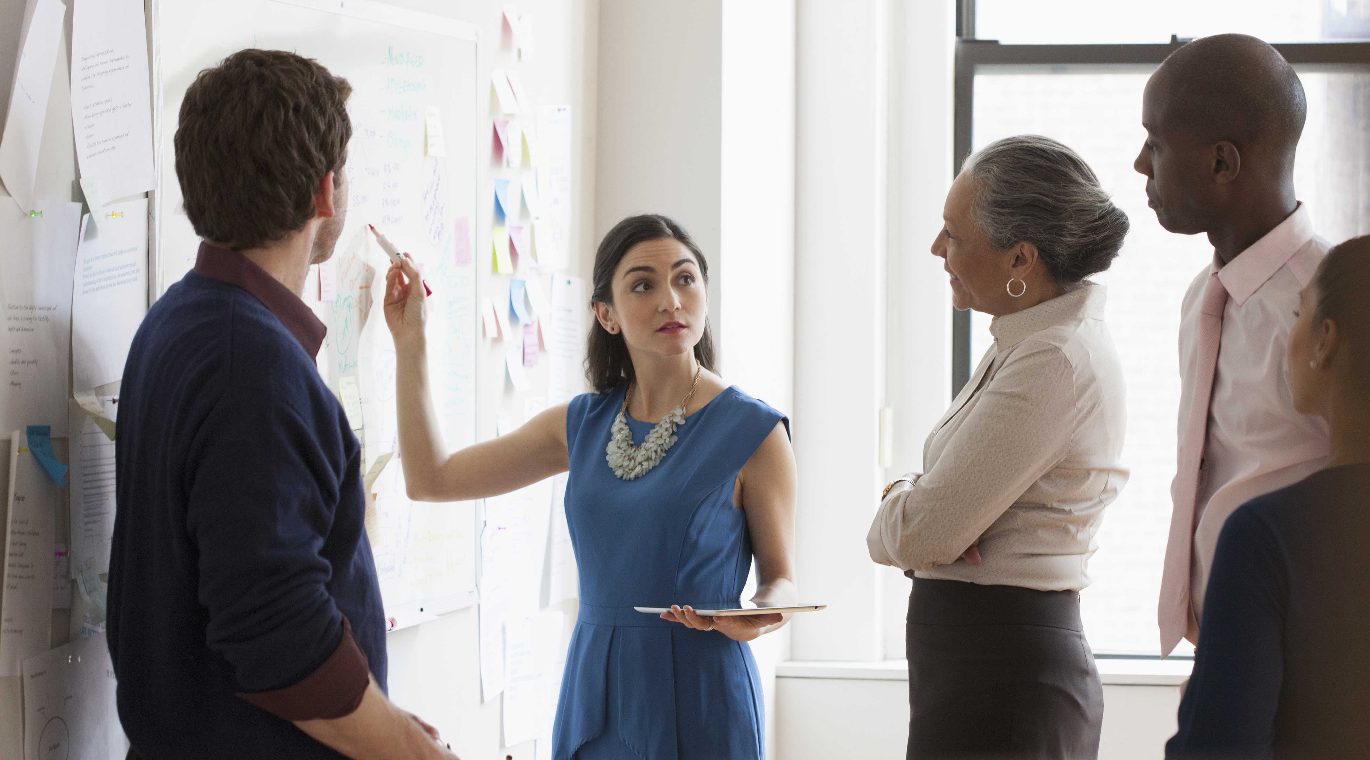 A group of people look at a woman as she writes something on a sheet of paper attached to a wall.