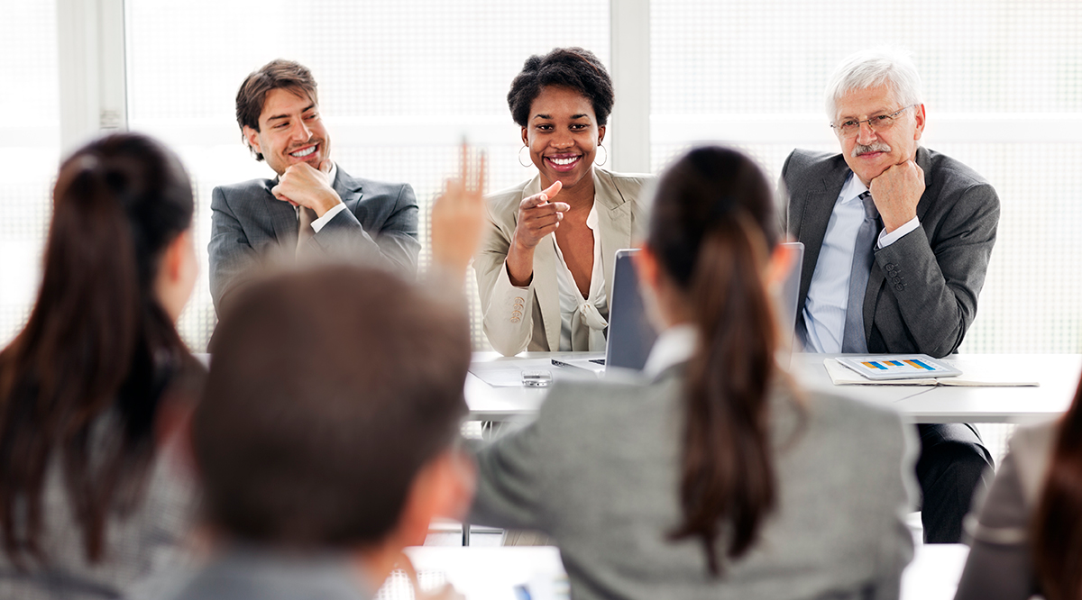 A woman takes questions at a meeting while two male colleagues look on supportively.