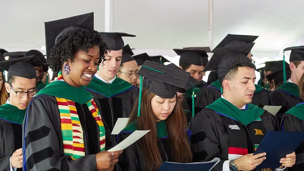 A group of medical school students are standing up, dressed in graduation caps and gowns, and reciting a medical oath. The photo focuses on four students.