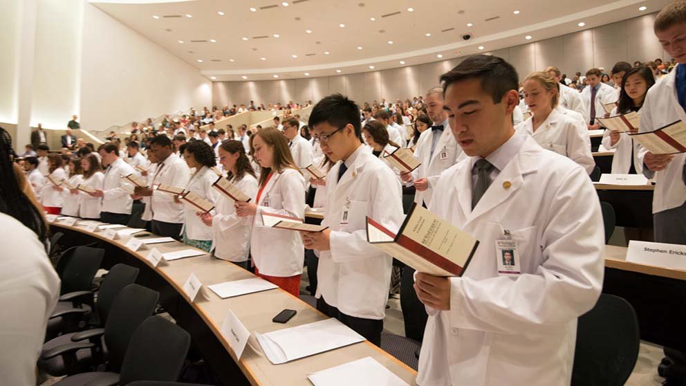 Medical students wearing white physicians’ coats stand in a large room. They read a printed oath out loud for their white coat ceremony.