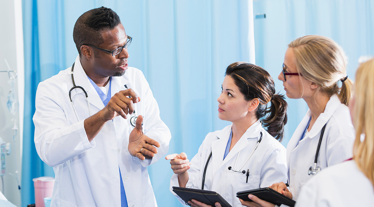 A group of multi-ethnic doctors in a hospital room, wearing white lab coats, having a discussion. Two of the female doctors are holding digital tablets.