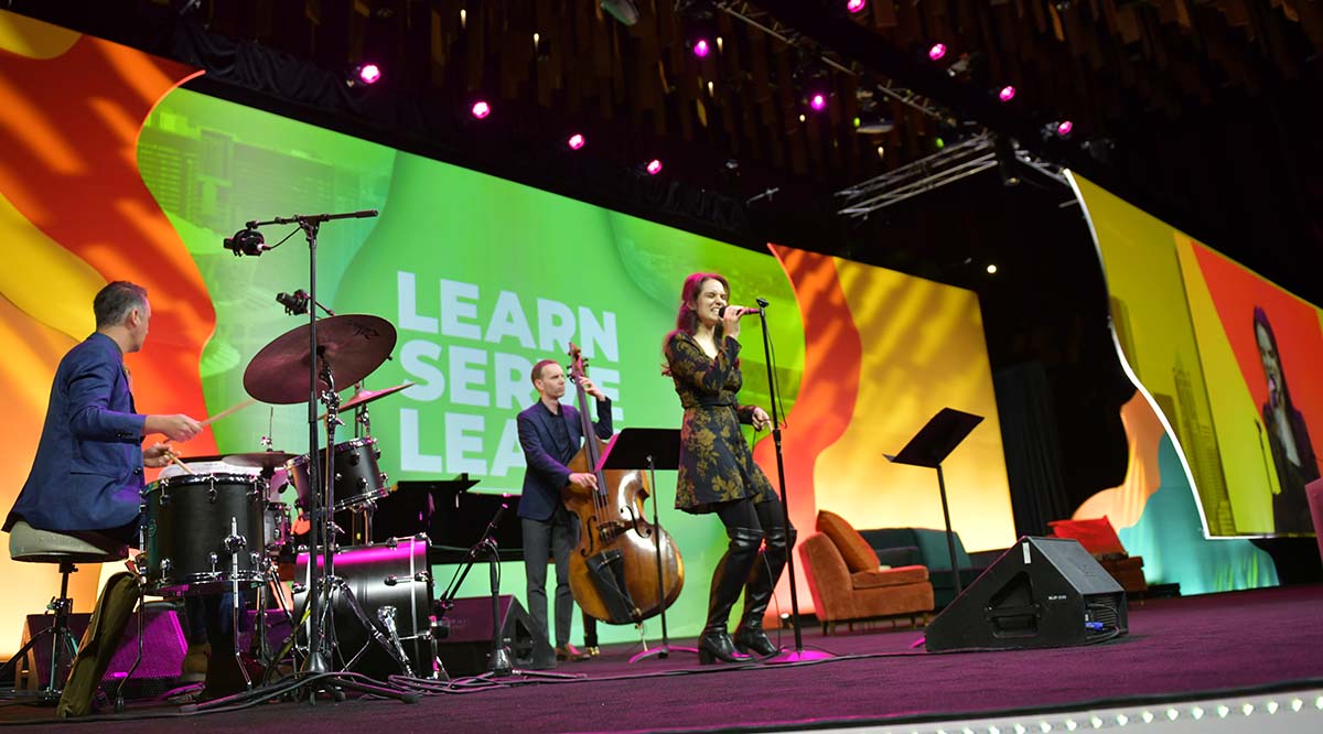 Three musicians -- a man on drums, a man on the standing bass, and a women singer -- perform on stage during Learn Serve Lead 2023.