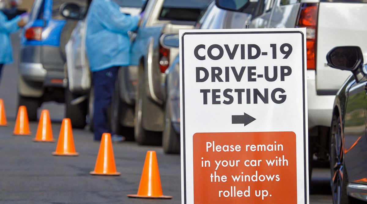 Health care workers perform COVID-19 tests along a line of cars. A sign says "COVID-19 Drive-Up Testing."