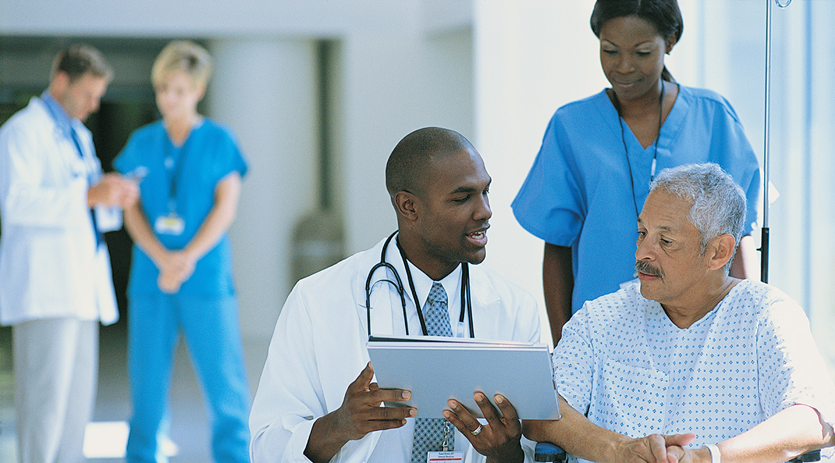 A doctor explains paperwork to a patient in a wheelchair while a nurse pushes him.