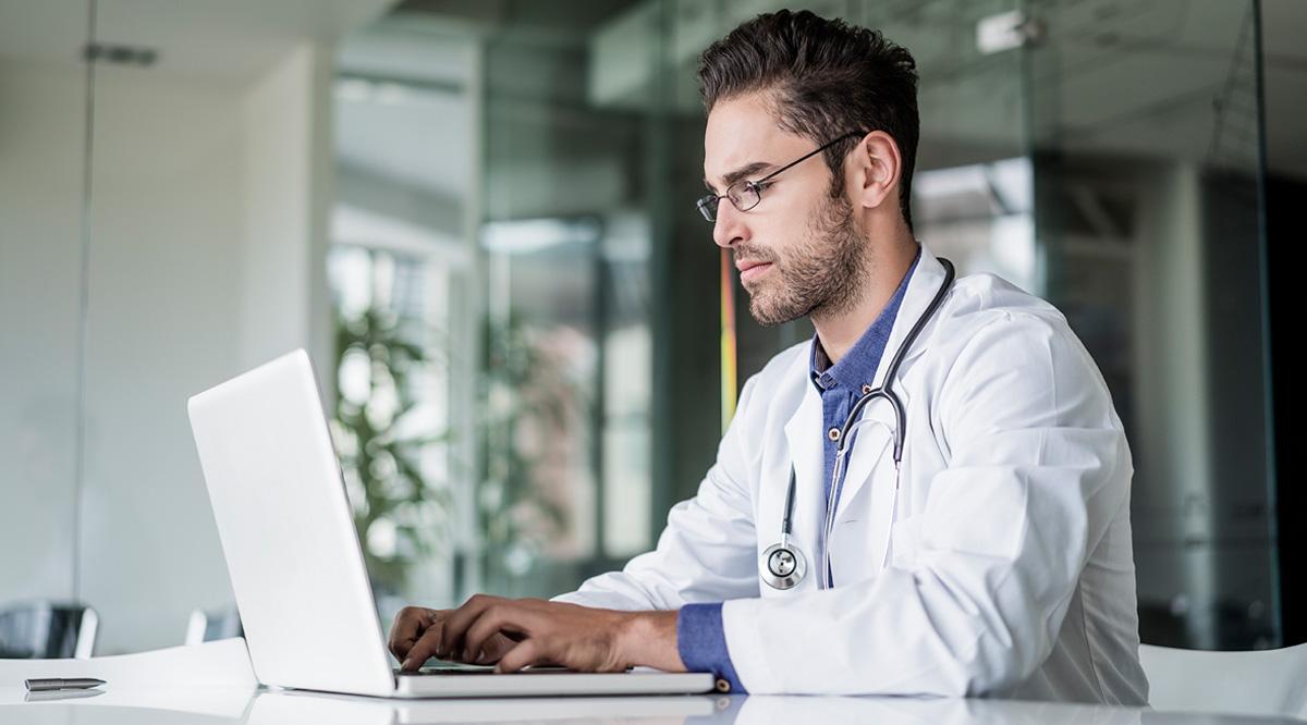 A male doctor, wearing a white coat and with a stethoscope around his neck, sits and works on a laptop computer.