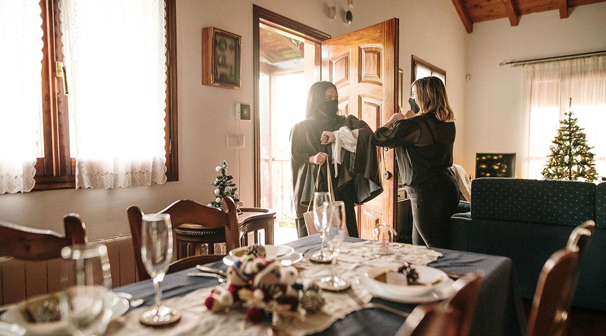 Two women in masks greet each other with an elbow bump in a house decorated for Christmas