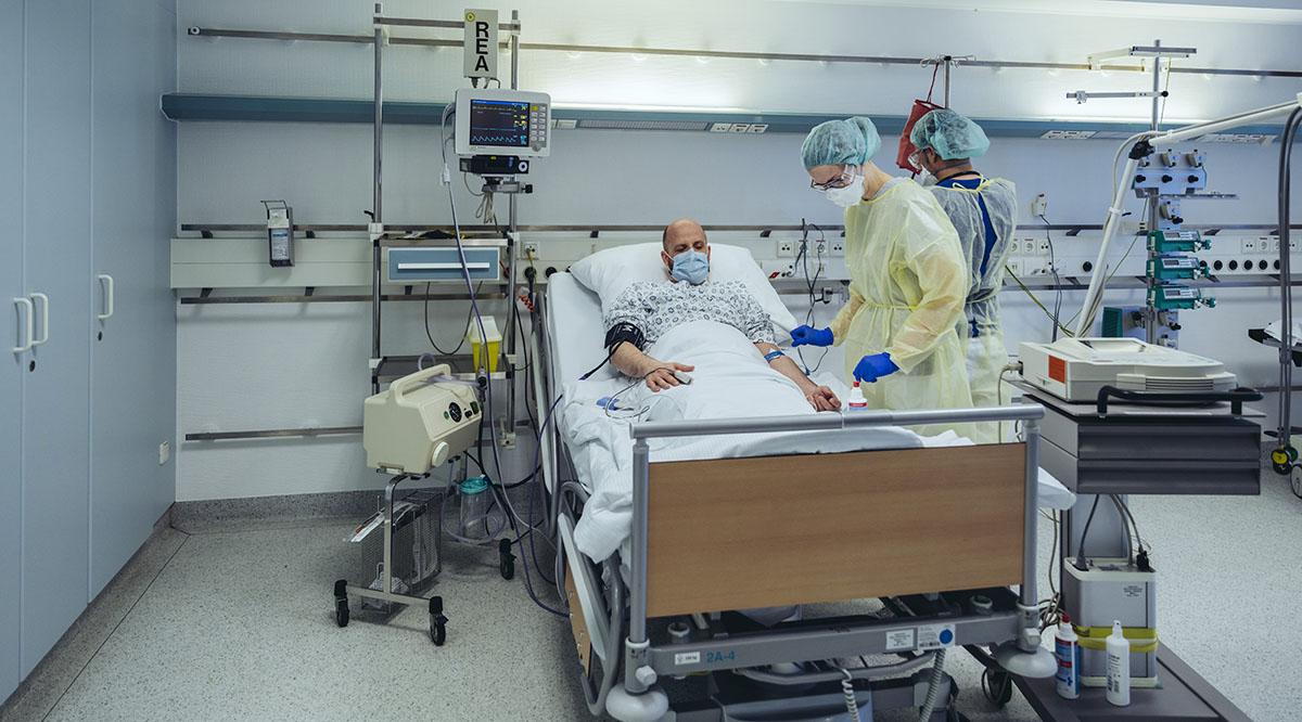 A patient in a mask is being cared for in the emergency room by two doctors in PPE