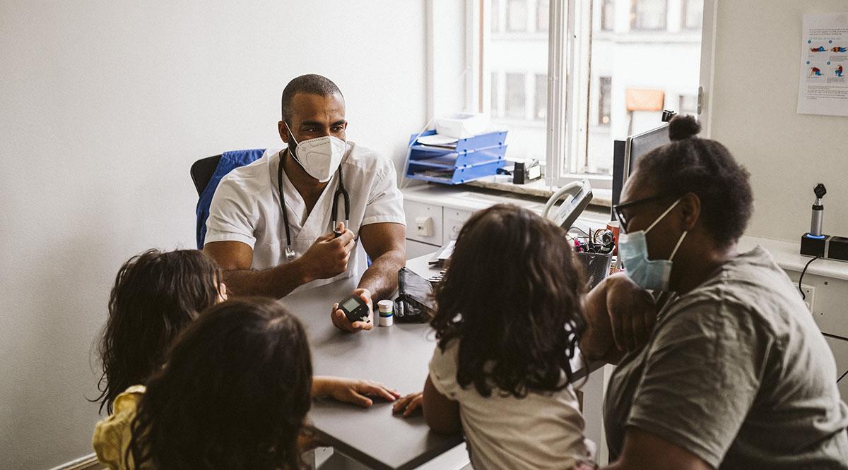 A masked doctor talks with a parent and children in his office