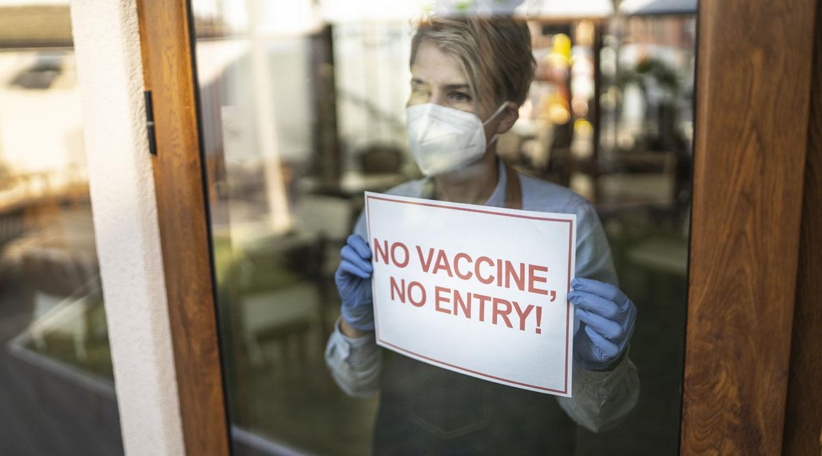 A masked woman stands near a sign that reads "No Vaccine, No Entry"
