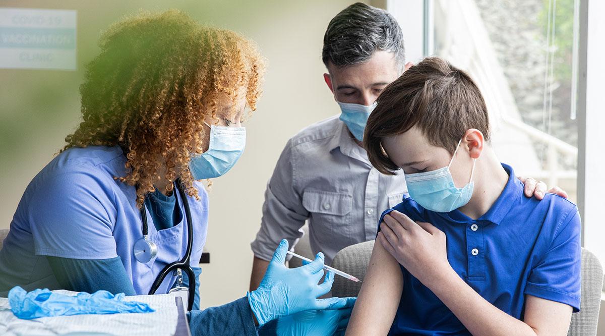 A masked teenager receives a vaccine from a masked provider
