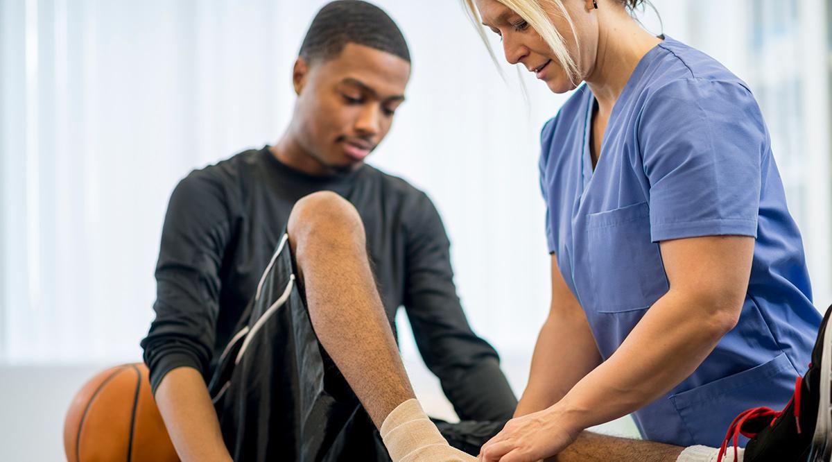 A male teenage basketball player is at the doctors office to have some work done on his injury. A nurse is wrapping his bandage around the ankle.