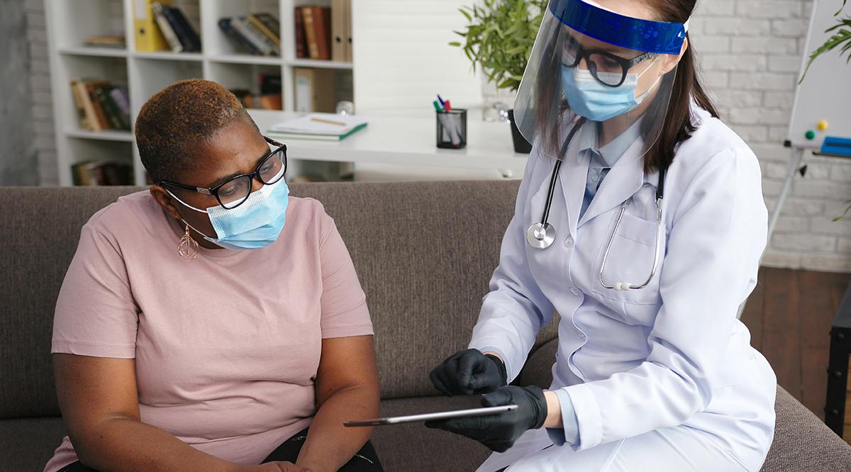 Doctor in protective equipment talks to obese African-American mature lady holding black tablet