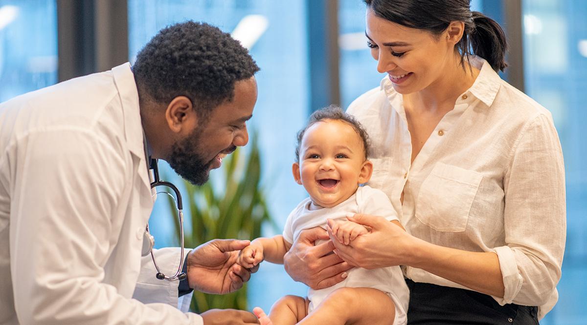 A mother brings in her son for a wellness check-up. She has the baby sitting on her lap with her hands firmly around his waist. The male doctor is reaching out to hold the baby's hand. The doctor is laughing and engaging the young boy.