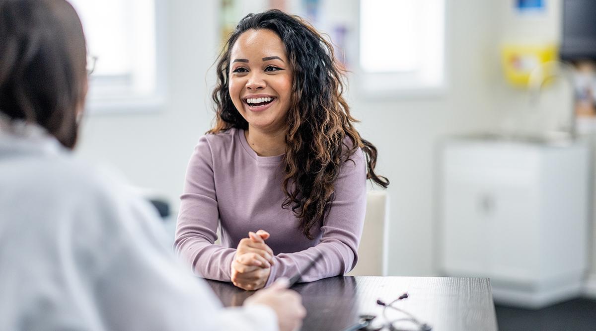 A young woman of mixed race, sits with her female doctor as they discuss her health concerns. The patient is dressed casually and has a smile on her face.