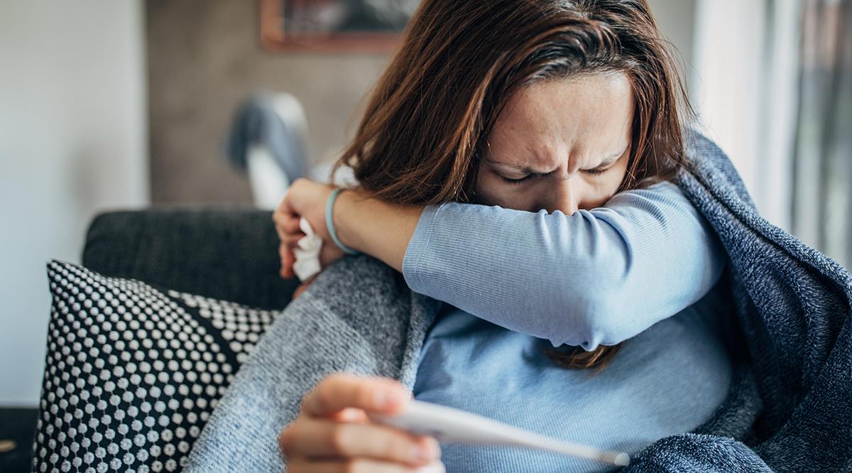 Woman with fever symptoms sitting on sofa and holding thermometer