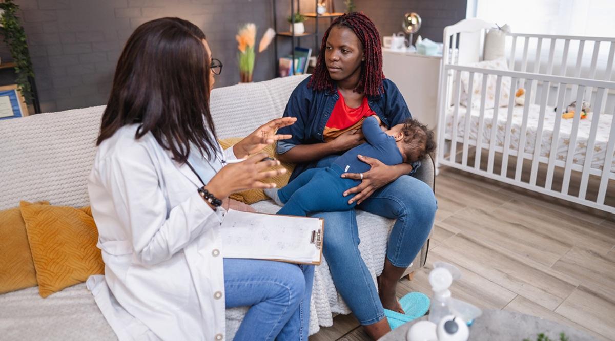 Doctor is visiting a young mother breastfeeding her baby at home, providing support and medical advice