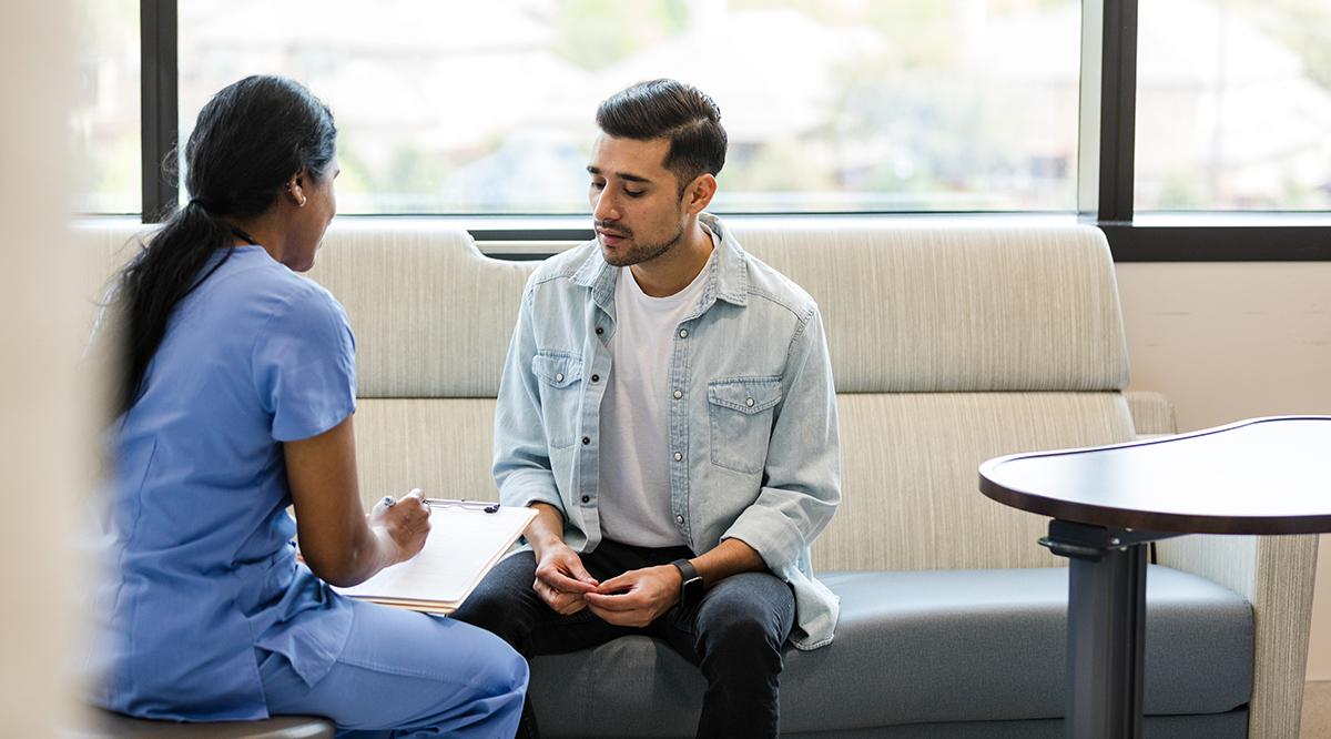 The young adult male sits on the hospital room couch to talk with the hospital insurance specialist to update medical information.