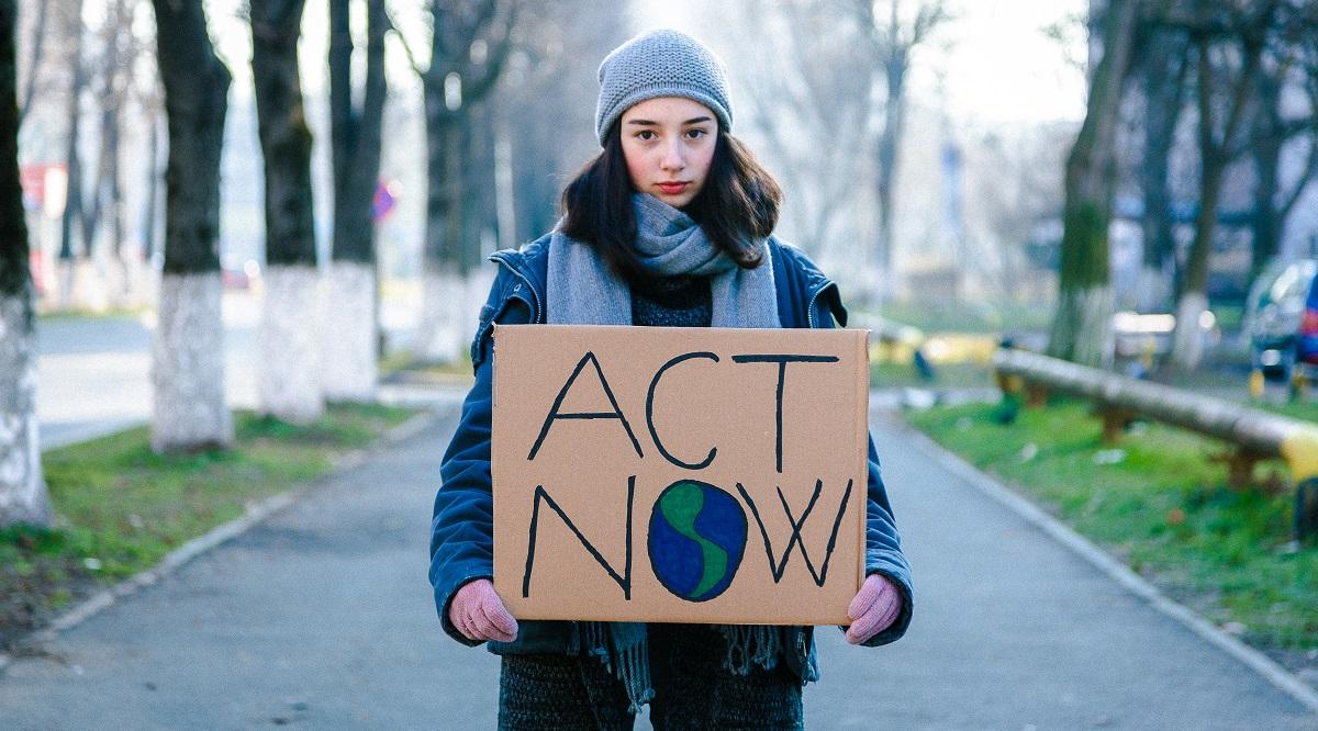 Portrait of a young teenage girl activist holding a sign protesting against climate change and global warming. She is standing outdoors on a city street and is holding signs with various messages, such as 'go green' and 'save our planet'. She is looking directly at the camera with a serious expression. Focus on the girl and her sign, with city street defocused beyond. Room for copy space.