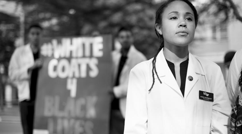 A woman in a white coat states in front of a sign that reads White Coats 4 Black Lives