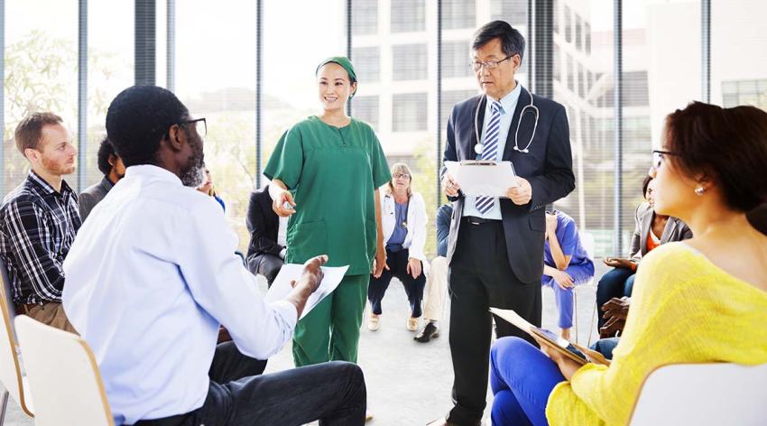 A male doctor wearing a suit and a stethoscope around his neck talks to a small group of people while a female medical worker wearing green scrubs stands next to him. 