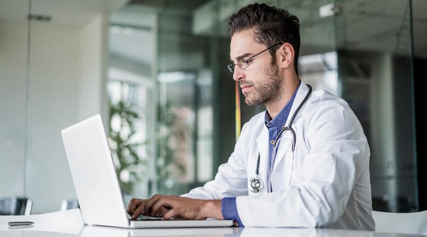 A male doctor, wearing a white coat and with a stethoscope around his neck, sits and works on a laptop computer.
