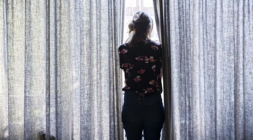 A woman stands alone looking out a window with curtains