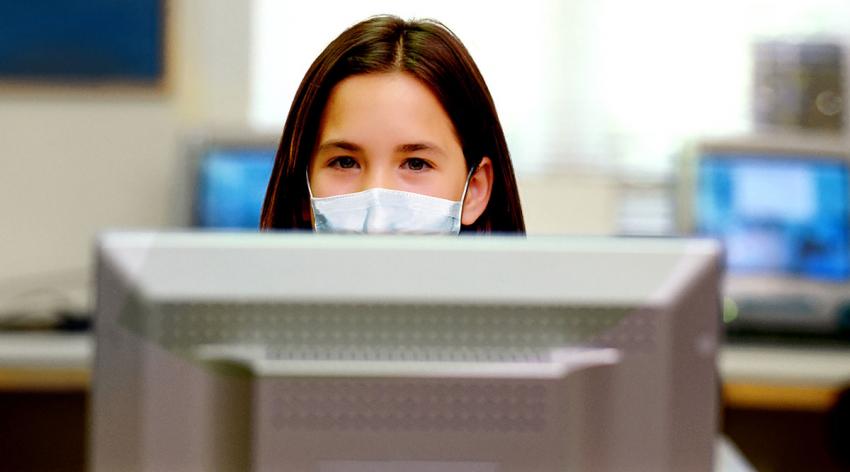 A woman in a mask sits at a computer taking an exam