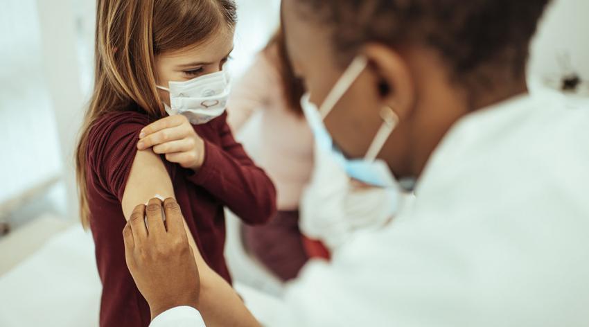 A masked doctor prepares a masked child patient for a vaccination