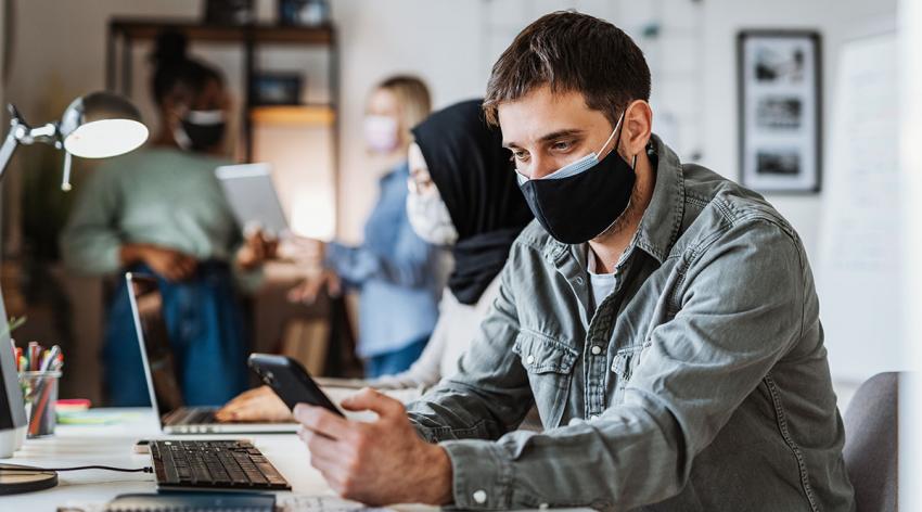 A double-masked office worker sits at a desk and looks at his phone