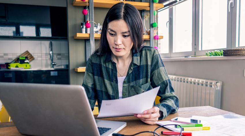 A woman looks intently at a laptop while holding a piece of paper