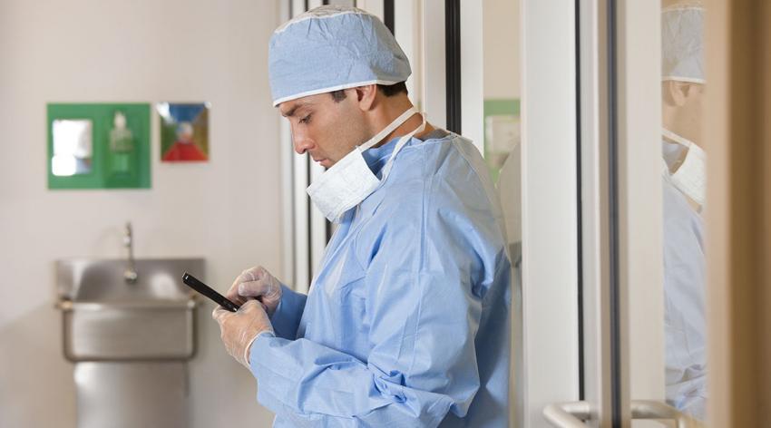 A doctor in scrubs leans against a wall and looks at his phone