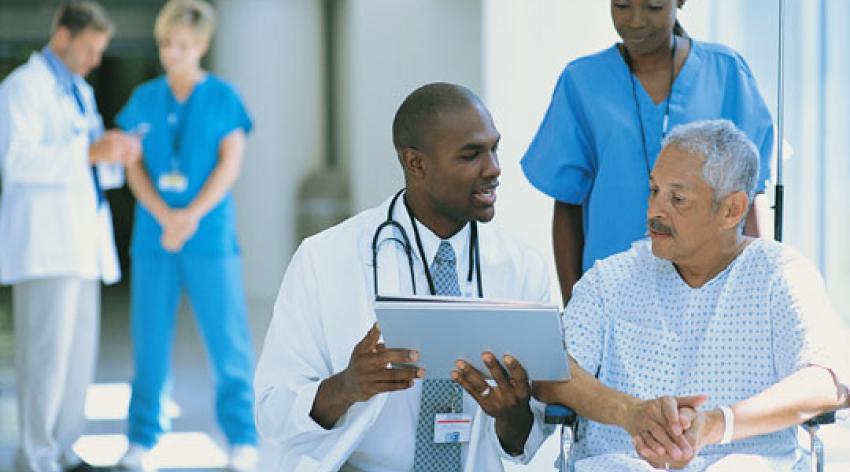 A doctor explains paperwork to a patient in a wheelchair while a nurse pushes him.