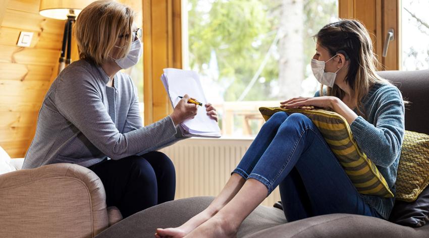 A young woman with a cochlear implant sits on a couch talking to her therapeutic practitioner.