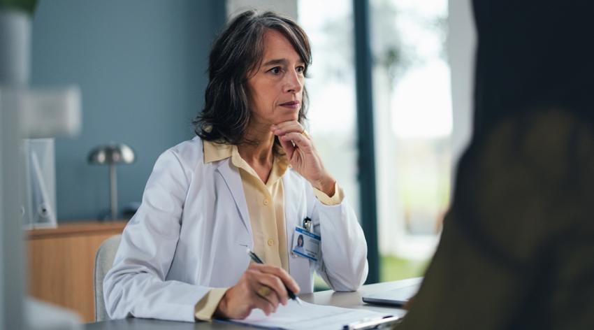 A thoughtful female doctor in a white coat sits at a desk with a clipboard, discussing patient details. The calm medical setting conveys professionalism, care, and focus in a clinical consultation.
