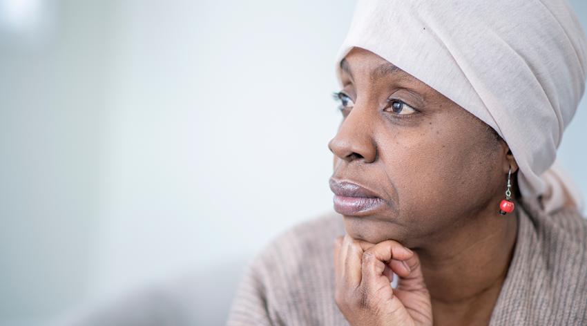 A mature African woman sits on a chair in the comfort of her home as she poses for a portrait. She is dressed comfortably in a light shirt with a brown shall thrown over her shoulders. She has a head scarf on and a neutral expression on her face as she holds her head up with her hand.