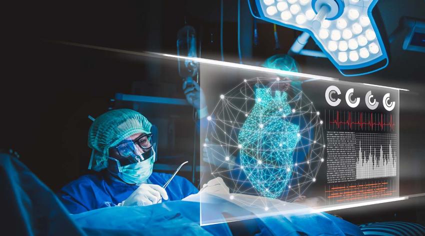 A doctor wearing blue scrubs and a mask looks at a digital video screen while performing surgery. 
