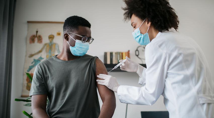 Young African American male patient sitting in a medical clinic and is being given the COVID-19 vaccine in his arm by a female African American doctor, both wearing protective face masks