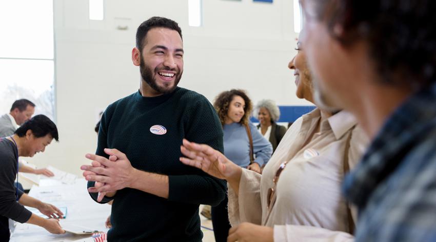 Smiling mid adult Hispanic man smiles and laughs with friends as they wait to register to vote.