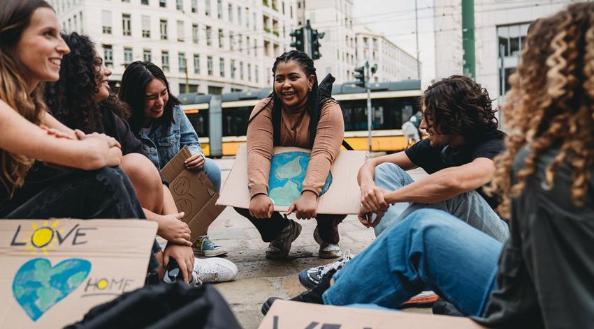 A group of young adult people are resting after a climate change demonstration in the city. They are sitting in circle with some cardboard signs against global warming.