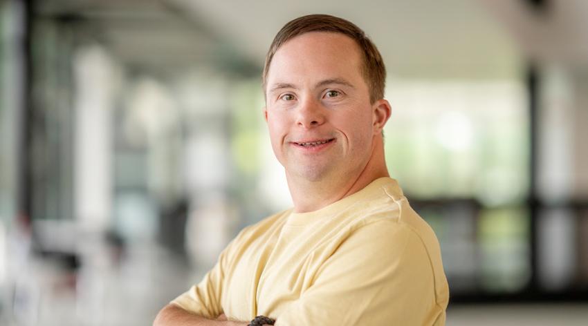 A man in his 30s smiles while standing with his arms crossed in a large, modern space.