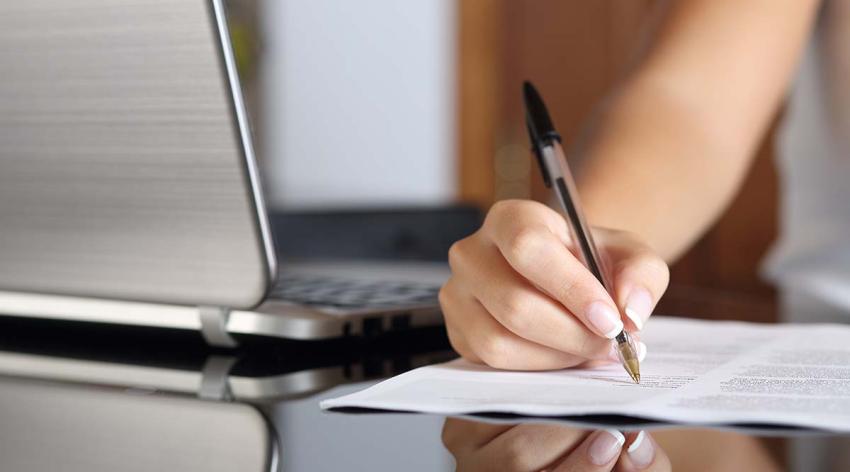 A person writes on a piece of paper next to a laptop. Only the person's arm and hand is in the photo. 