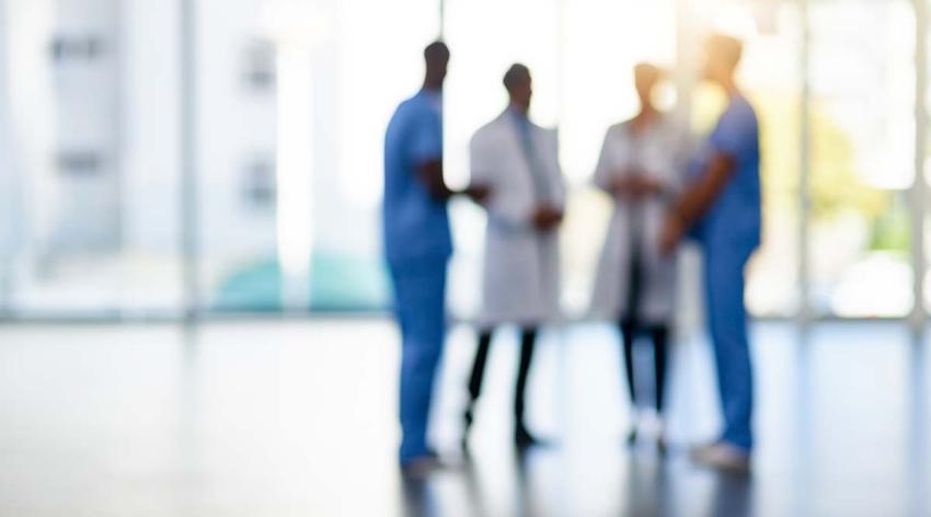 Four medical workers are seen talking in a room with a large window behind them. The four people are blurred, but you can see that two are wearing scrubs and two are wearing white coats.