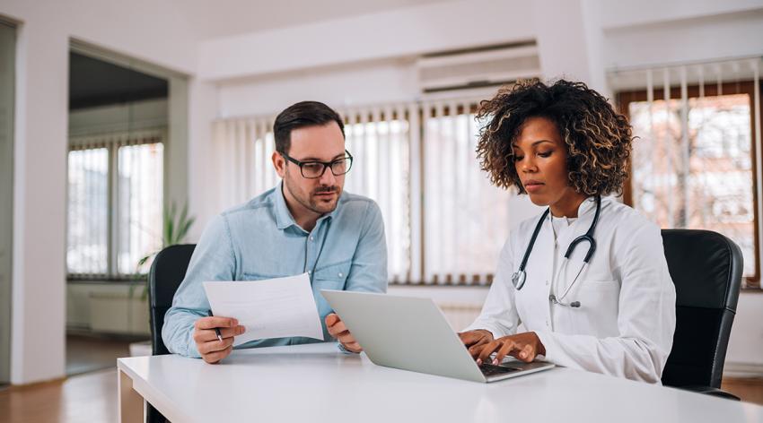 Female doctor, resident, using a laptop while sitting at desk with mentor who looking and holding paper document.