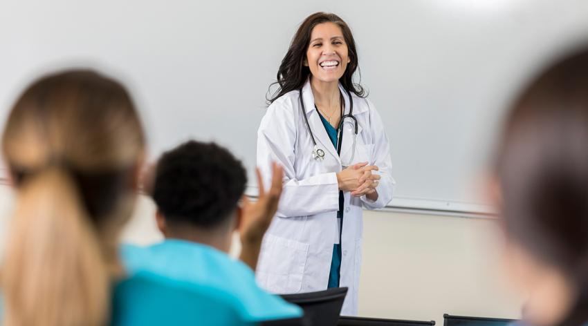 A female medical professor stands in front of her classroom and laughs at a student as he adds humor to her lecture.