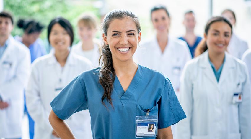 A female medical student of Hispanic ethnicity smiles for the camera as she stands in front of her group of medical professionals in the background.