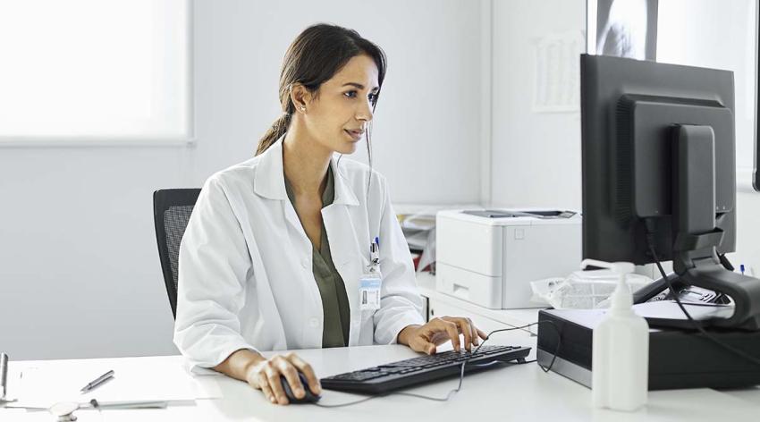 A female doctor sits at her desk wearing a white coat while looking at a computer monitor and typing. 