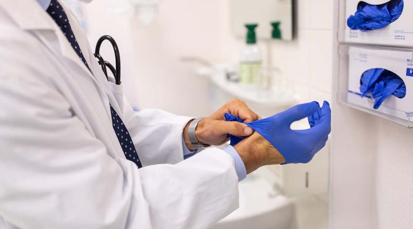 A doctor wearing a white coach, whose face cannot be seen, puts on blue gloves.
