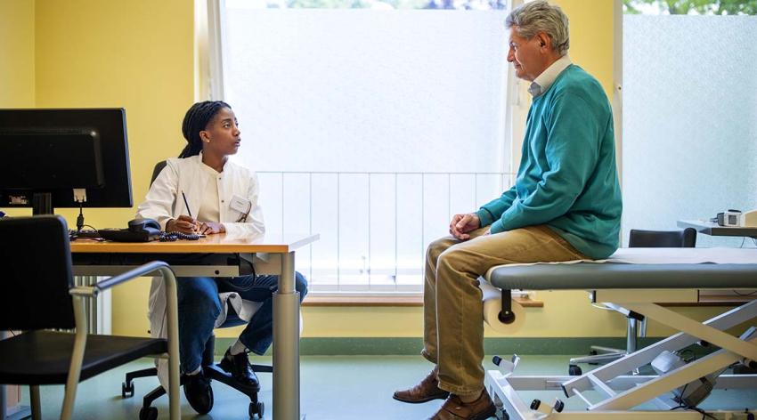 A female doctor sitting at a desk with a computer on it talks to a male patient sitting on an examination table.